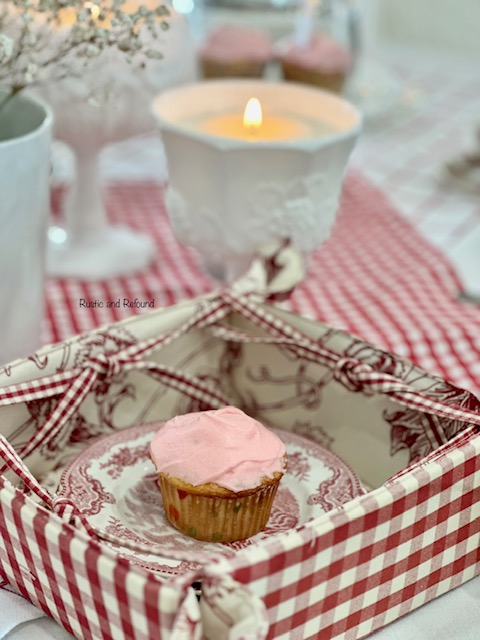 A pink cupcake on red dessert plates with a candle in a milk glass compote.